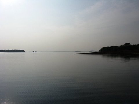 A wide expanse of flat, silvery water under a hazy sky, with a wooded shoreline on the right and distant islands fading into the mist.