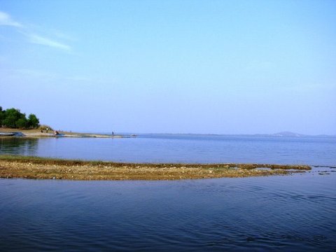 A low sandbar curves across deep blue water beneath a clear sky, with boats and trees on the far shore to the left.