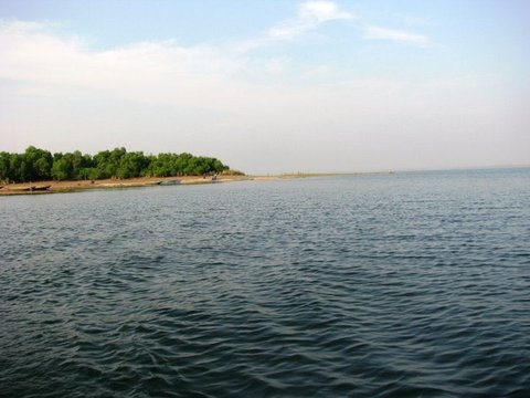 Choppy blue-green water in the foreground with a tree-lined shore and a few beached boats under a pale sky.