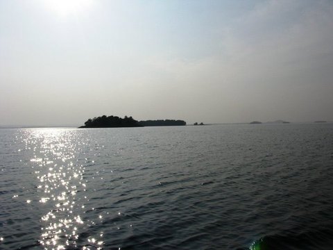 Sunlight glitters in a path across rippled water towards a dark wooded island, with smaller islands fading into the haze beyond.