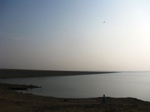 A long straight embankment lines the far edge of a calm reservoir under a hazy sky, a beached boat and a lone figure on the bare shore.