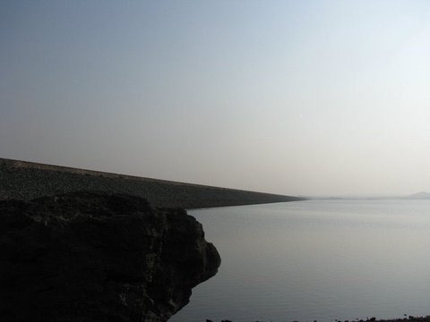 A dark boulder in the foreground with a long stone-pitched embankment stretching away beside flat, pale water under a hazy sky.