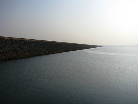 A stone-faced embankment runs in a straight diagonal into the distance, its base meeting calm grey water beneath a pale sky.