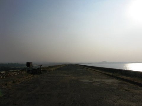 An empty paved road runs straight along the top of a dam into the haze, trees on the left and sunlit water on the right.