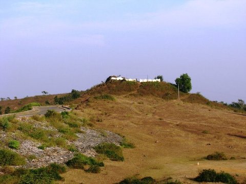 A dry grassy hill with scattered bushes and a small white building on its crest, a narrow road curving round its base.