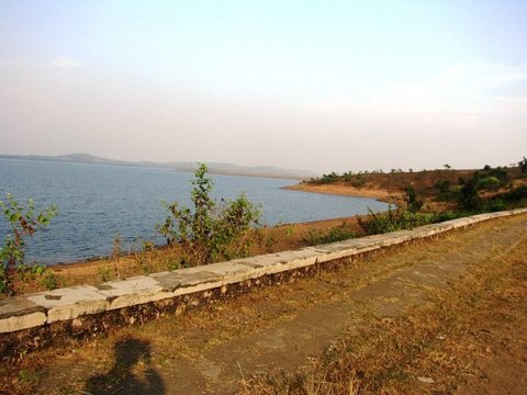 A low whitewashed stone wall and dirt track curve along a reservoir shore, dry grass in front and low hills across the pale blue water.