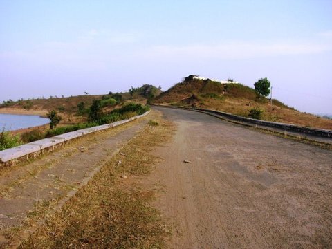 An empty tarmac road runs along a grassy embankment toward a scrub-covered hillock, water visible to the left beneath a hazy blue sky.