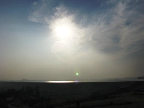 The sun burns through a wisp of cloud above a distant strip of water, dark scrubland in silhouette along the bottom of the frame.