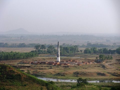 A tall tapering brick chimney rises from a kiln surrounded by stacked red bricks, with fields, trees and a hazy hill beyond.