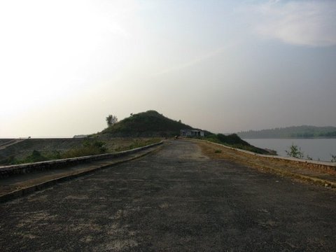 A worn tarmac road leads toward a grassy hillock with a small white hut at its foot, calm grey water to the right under a pale sky.