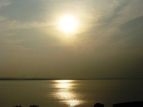 A hazy golden sun hangs above a flat reservoir, its reflection a bright shimmering path across the water toward a dark shoreline.
