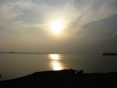 A soft sun glows through haze above a wide reservoir, with a wooded island at right and a low hill on the far horizon.