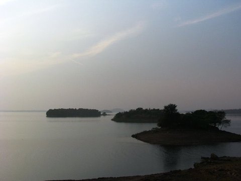 Two small wooded islands sit in flat grey water under a pale hazy sky, a tree-topped spit of land reaching in from the right.