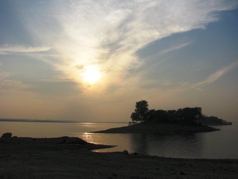 The sun glows through streaked cloud above a small tree-covered island, its light reflected on the water beside a dark rocky shore.