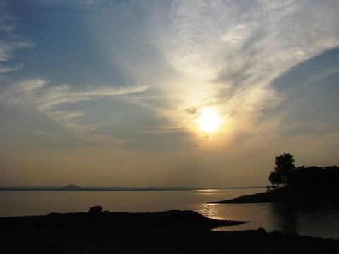 Streaks of cirrus cloud fan across a blue sky around a low sun, with a dark shoreline, a lone tree and a hill across the water.