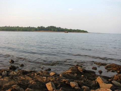 Rippled water laps against a rocky shore, with a small boat passing in front of a long wooded bank across the reservoir.