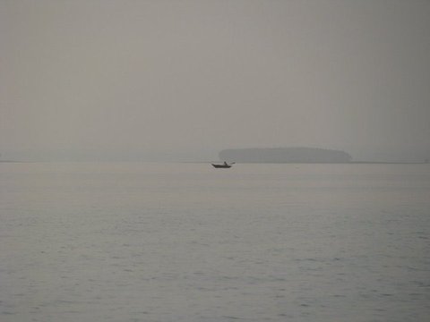 A small boat with a single figure drifts on flat grey water in thick haze, a wooded island faint on the horizon behind it.