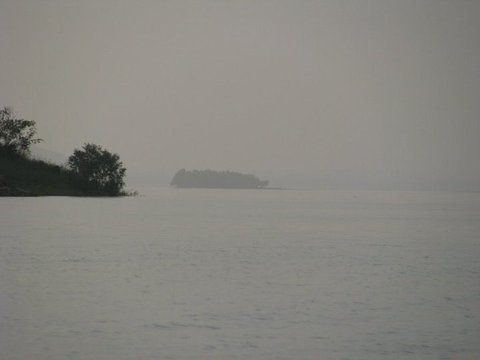 A wooded island dissolves into grey haze across still water, with scrubby trees on a near bank at the left edge.