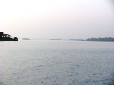 Rippled water stretches toward a line of low wooded islands and a tiny boat on the horizon beneath a pale, hazy sky.