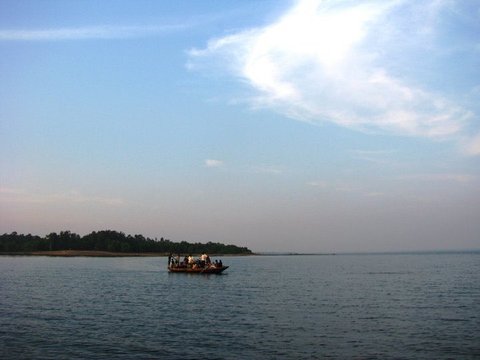 A small wooden boat carrying a group of passengers crosses calm blue water beneath a wide sky with a sweep of thin cloud.