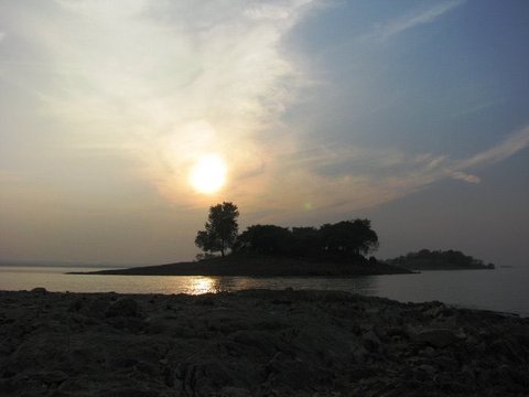 A dark rocky foreshore leads to a wooded island silhouetted against a hazy sun, its reflection glinting on the water.