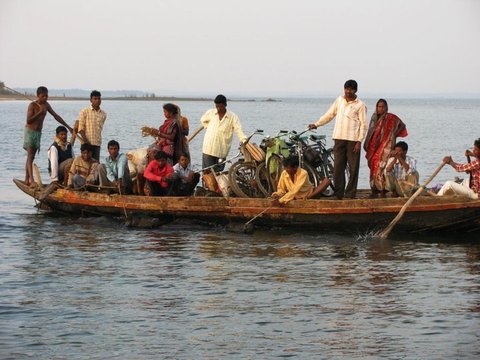 A crowded wooden ferry boat with bicycles stacked amidships is poled across calm grey water, passengers standing and seated along its length.