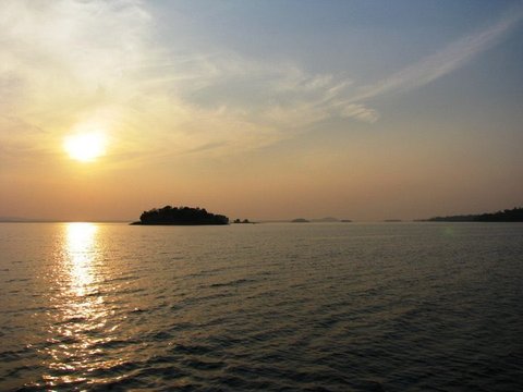 A hazy sun hangs above a small wooded island, laying a golden track across gently rippled water as distant islets fade into the horizon.