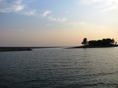 Rippled blue water leads to a low wooded island at right, its trees silhouetted against a pale evening sky.