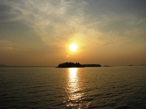 A golden sun glows through haze above a dark wooded island, its reflection a shimmering column on the water below.