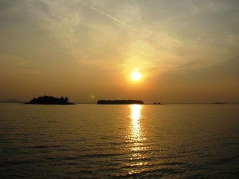 The low sun casts a golden path across rippled water past two silhouetted islands, a thin contrail crossing the amber sky.