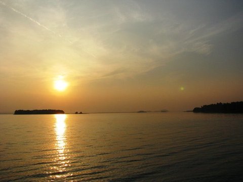 A hazy sun above a low wooded island throws a bright reflection across calm water, with tree-lined shores fading at either side.