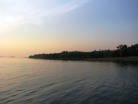 A low wooded shoreline darkens against a pale orange and blue sky, its reflection trembling on the rippled surface of the reservoir.