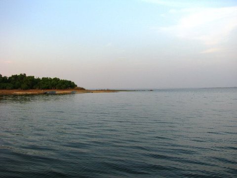 A tree-covered point of land tapers into the reservoir on the left, a boat moored at its tip, with hazy sky over the wide grey water.