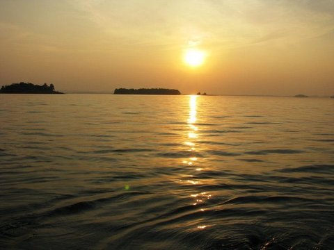 A golden sun hangs above dark wooded islands, its reflection a bright ribbon across the rippling reservoir.
