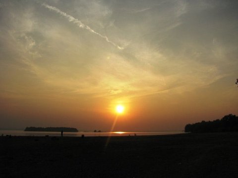 The setting sun glows amber beneath streaked cloud, its reflection reaching across the water toward a dark beach where a lone figure stands.