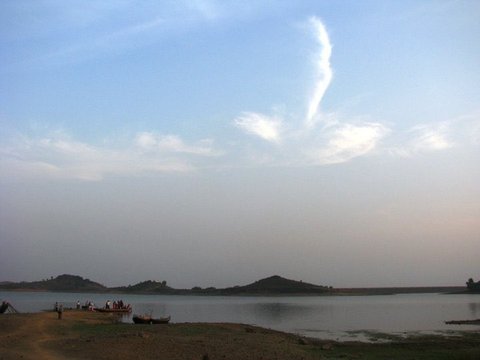 Small boats and a cluster of visitors gather on a muddy bank beneath two low hills, a single wisp of cloud rising in a pale blue sky.