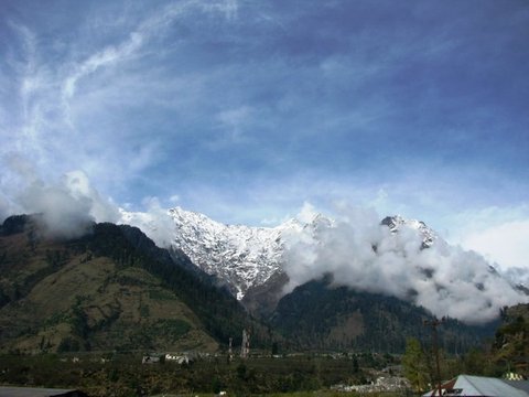 Snow-covered peaks rise behind a forested ridge, cloud gathering at the summits beneath a blue sky streaked with cirrus.