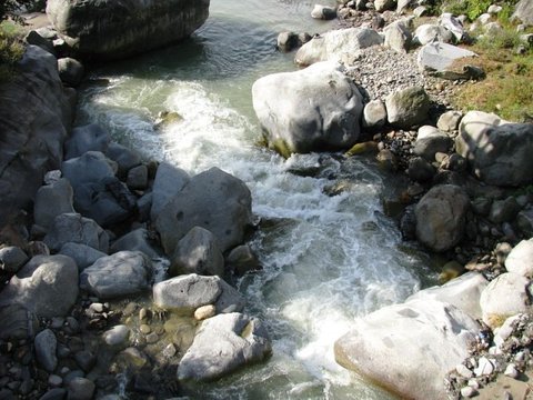 A glacial-green stream churns white over rounded grey boulders, seen from above in strong sunlight.