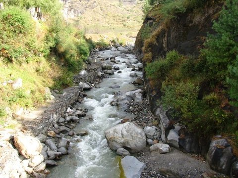 A fast, pale-green stream threads boulders between a dark rock wall and a stone-walled path lined with shrubs.