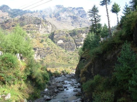 Tall pines on a rocky bluff overlook a boulder-strewn stream, with terraced cliffs and a hazy mountain wall behind.