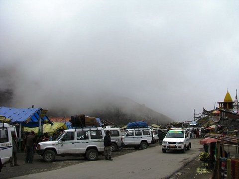 White SUVs with luggage on their roofs wait on a muddy road beside tarpaulin stalls as mist hides the mountain above.