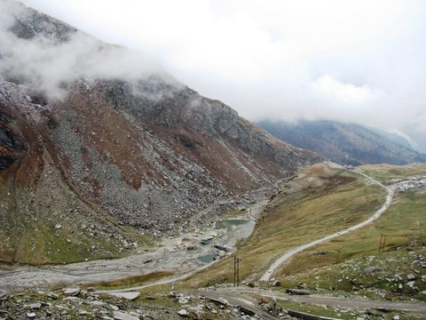 A grey, snow-dusted scree slope descends to a small green pool and a gravel road winding across moorland beneath low cloud.