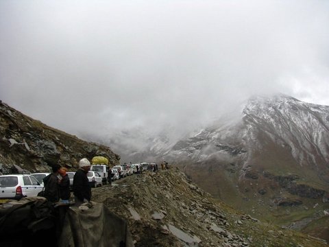 A line of white vehicles waits on a gravel road beneath a snow-dusted ridge wrapped in grey cloud, a few people at the roadside.