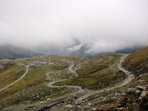 A gravel road zigzags across green slopes past a huddle of roadside stalls while cloud fills the valley beyond.