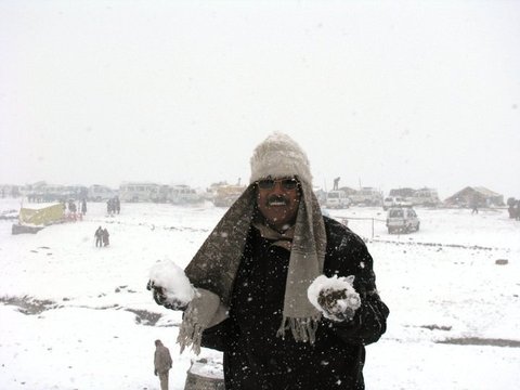 A man in a dark coat, knitted hat and shawl holds handfuls of snow as flakes fall, parked vehicles and tents blurred behind him.