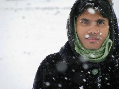Close portrait of a man in a dark hooded coat and green scarf, snowflakes drifting in front of a white, snow-covered ground.