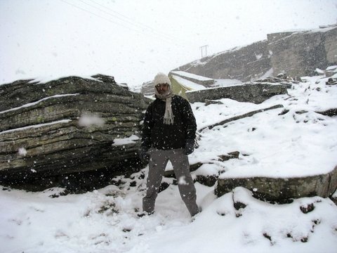 A man in a black jacket, woollen cap and scarf stands among snow-covered boulders as thick flakes fall against a white sky.