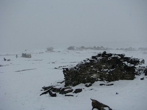 Falling snow blurs a line of low huts on a white plateau, with a tumbled dry-stone wall buried in fresh snow in the foreground.