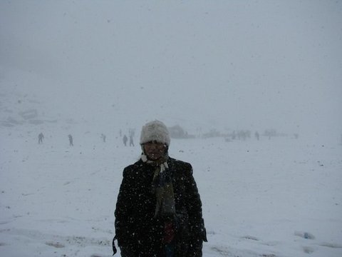 A person in a white cap and dark coat stands in heavy snowfall, small figures and huts barely visible on the white plain behind.