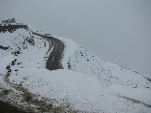 A lone vehicle rounds a dark hairpin bend cut through deep fresh snow, more vehicles waiting on the ledge above under a grey sky.
